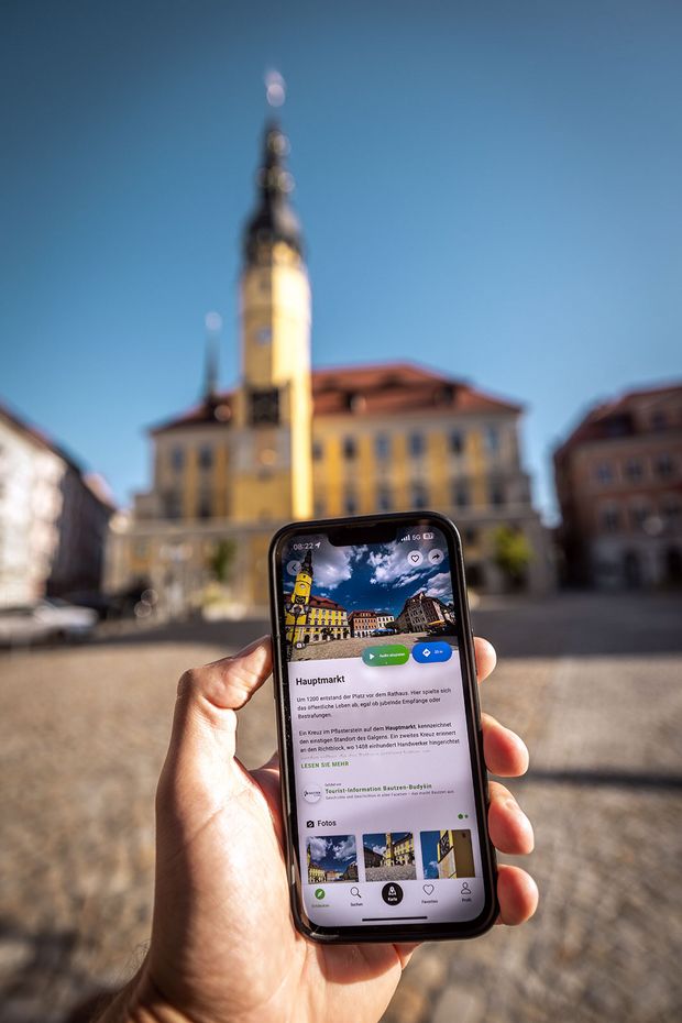 Eine Hand hält ein Handy in die Kamera, im Hintergrund ist ein großes gelbes Gebäude mit einem Turm zu sehen.
