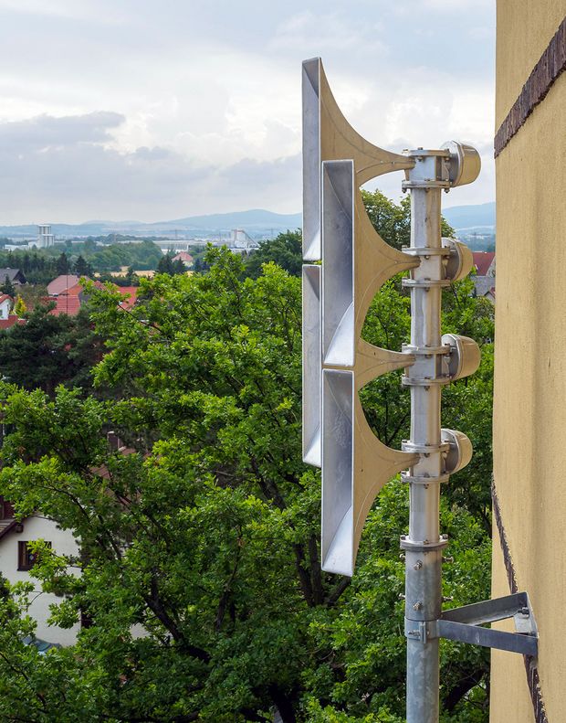 Rechts im Bild hängen gelbe Sirenen an einer gelben Hauswand, primär links und im Hintergrund ist ein grüner Wald sichtbar, der Himmel ist bewölkt