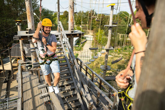 Kletterpark Vordergrund Person zu sehen, Hintergrund andere Person klettert über Balken