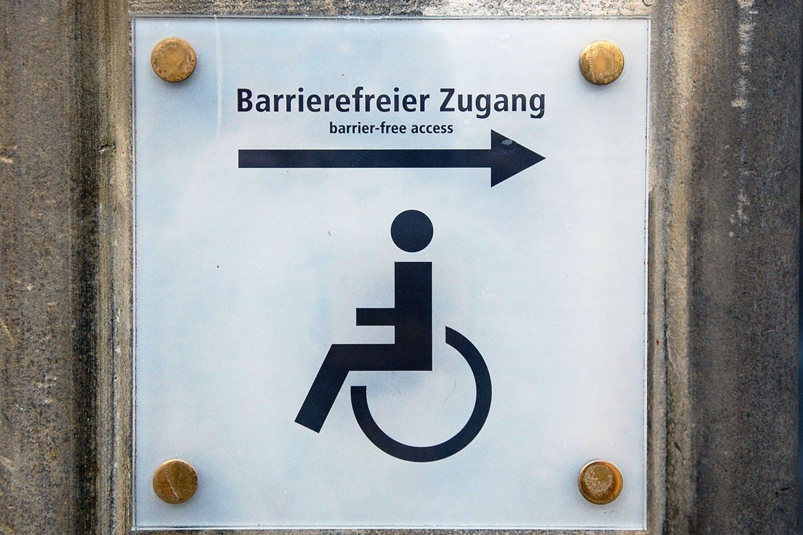 Weißes Schild mit Schriftzug barrierefreier Zugang darunter das Piktogramm für Rollstuhl
