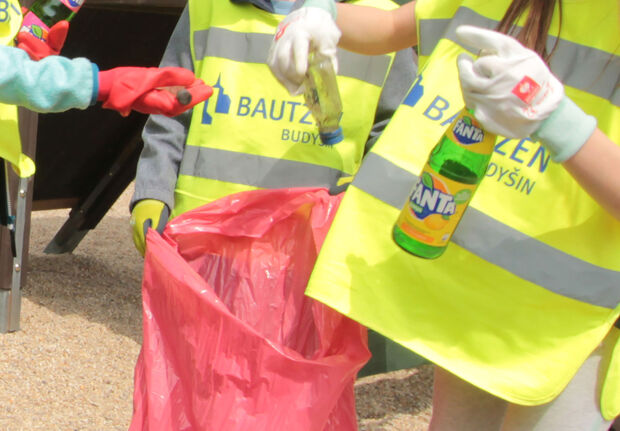 3 Personen beim Müllsammeln. Sie tragen gelbe Warnwesten mit dem Bautzen-Logo drauf und Handschuhe. Sie haben z.B. eine Fanta-Flasche. Eine Person hält eine große rote Mülltüte auf.