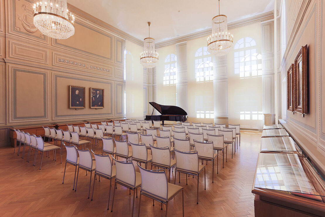 bestuhlter Saal, mit einem schwarzem Flügel, Deckenkronleuchtern, historischen Porträts an der Wand, großen Bogenfenstern und Vitrinen an der Seite