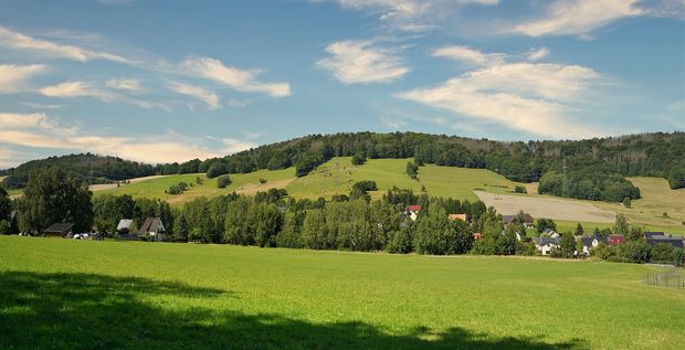 Großer Picho Grüne Wiese im Vordergrund dahinter ein bewaldeter kleiner Berg. Der Himmel ist blau mit kleinen weißen Schleierwolken.