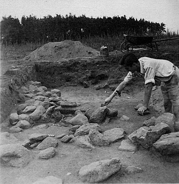 Schwarzweißfoto der Grabungen in den 50er Jahren in Niederkaina: zu sehen ist ein freigelegtes Grab mi keramischen Grabbeigaben und ein Mann der es gerade ausgräbt.