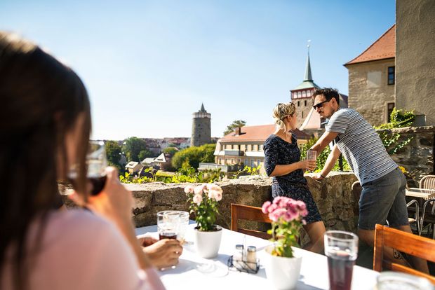 Auf Terrasse- an der Mauer stehen zwei Menschen und reden, nenben ihnen steht ein Tisch an welchen noch weitere Menschen etwas trinken; Aussicht auf Michaeliskirche und Alte Wasserkunst