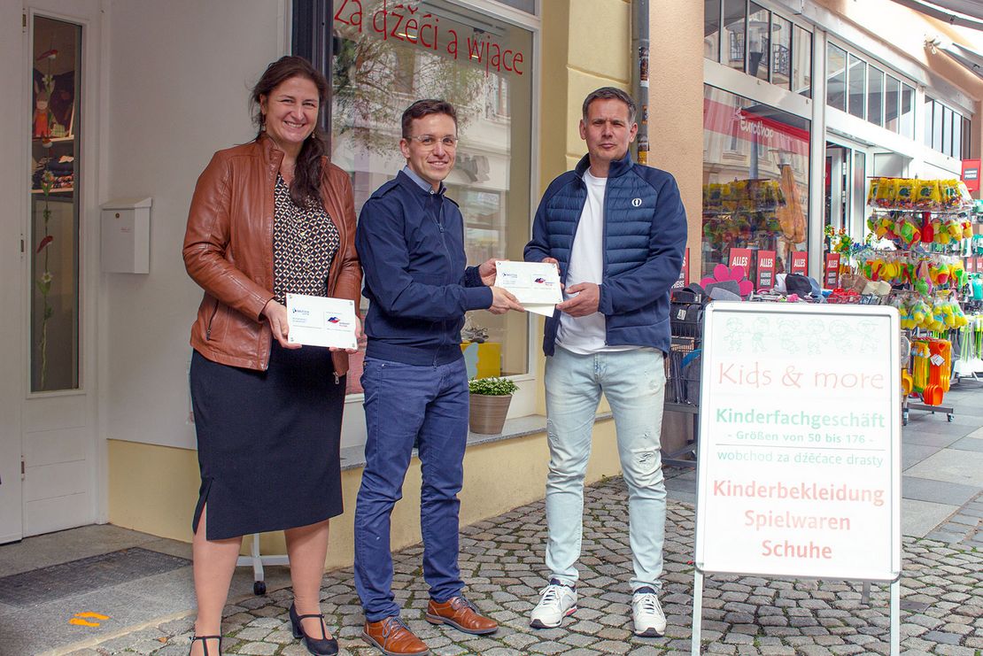 Eine Frau und zwei Männer stehen vor dem Geschäft kids and more und halten eine Plakate in der Hand