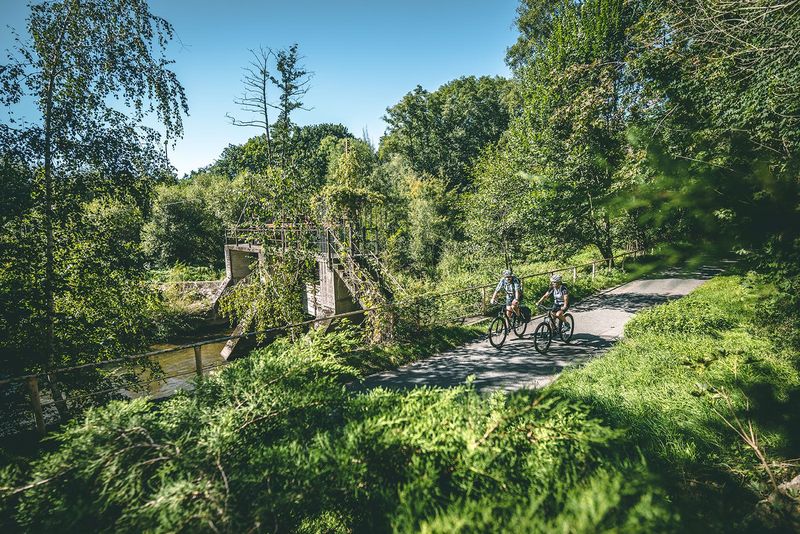 Zwei Radfahrer fahren auf einem Radweg vorbei an einem Wehr. Rechts und links stehen Bäume.