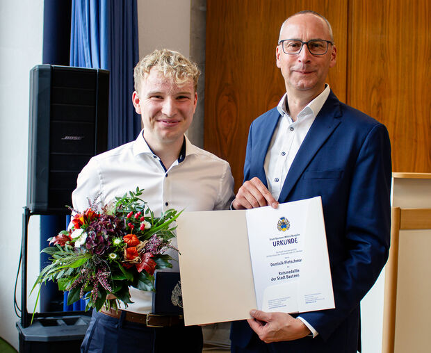 Junger Mann in weißem Hemd hält einen Blumenstrauß in der Hand. Daneben steht ein Mann mit weißem Hemd und blauen Jackett. Er hält eine Urkunde in der Hand.