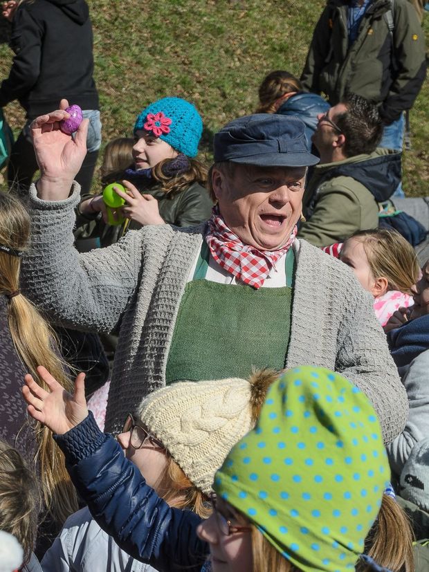 ein älterer Mann hält ein lila bemaltes Osterei in seiner rechten Hand nach oben, während Kinder vor ihm stehen