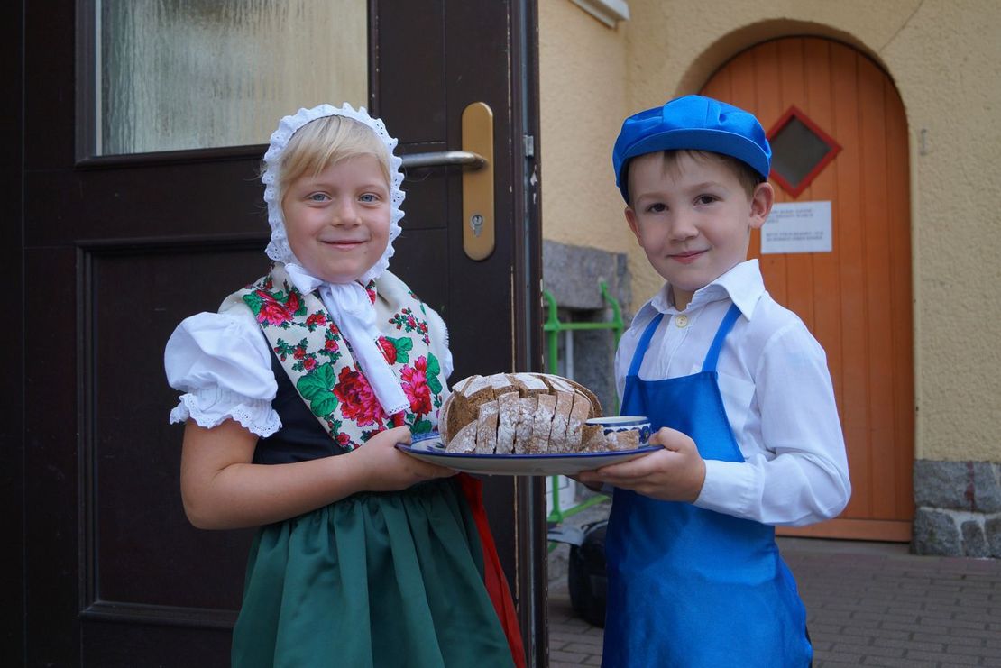 zwei Kinder (Junge und Mädchen) stehen in typisch sorbischer Tracht vor dem Eingang der Kita, und halten ein traditionelles Osterbrot in der Hand