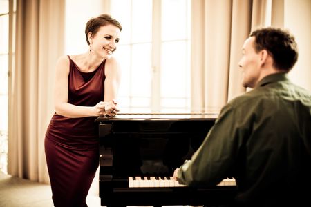 Foto von einem Mann am Klavier in Rückenansicht. Am Klavier lehnt eine lächelnde Frau in rotem Abendkleid mit kurzen, dunklen Haaren. Im Hintergrund seiht man ein mit beigen, halb durchsichtigen Vorhängen verhangenes Fenster.