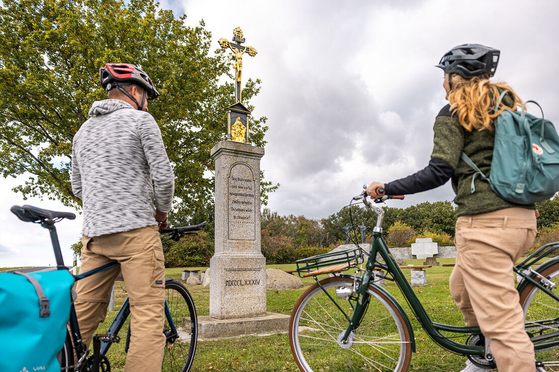 zwei Radler vor Steinsäule mit goldenem Kreuz