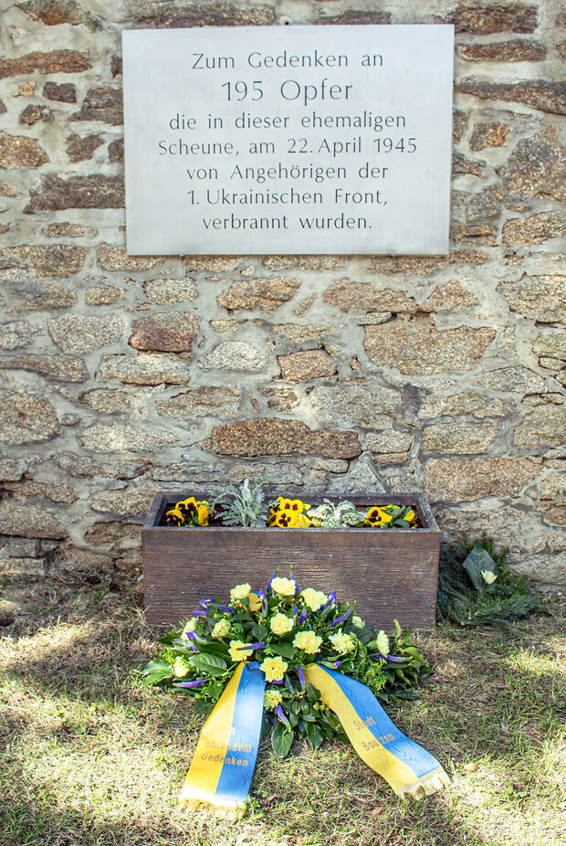 Blumenkranz vor Gedenktafel an Mauer. Aufschrift Tafel: Zum Gedenken an 195 Opfer die in dieser ehemaligen Scheune, am 22. April 1945 von Angehörigen der 1. Ukrainischen Front, verbrannt wurden.