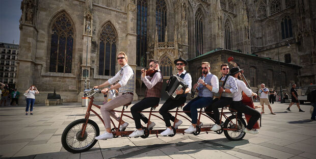 die sechs Musiker mit Instrumenten auf einem mehrsitzigen Fahrrad vor einer Kirche