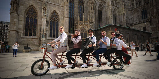 die sechs Musiker mit Instrumenten auf einem mehrsitzigen Fahrrad vor einer Kirche