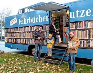 Bücherbus Blauer Bus mit Beklebung einer Bücherwand, davon stehen ein Mann mit einem Kind auf dem Arm, daneben ein Kind und eine Frau