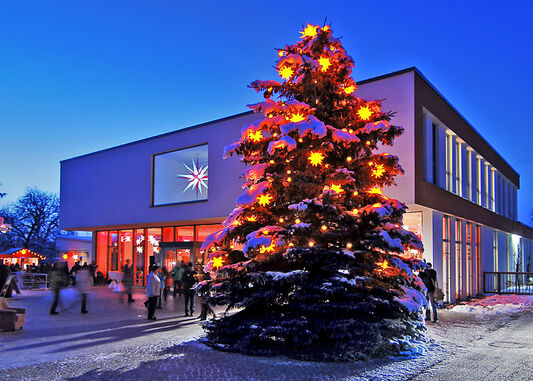 Herrnhuter Sternemanufaktur im Winter Im Vordergrund schneebedeckter Weihnachtbaum vor einem hell erleuchteten Gebäude.