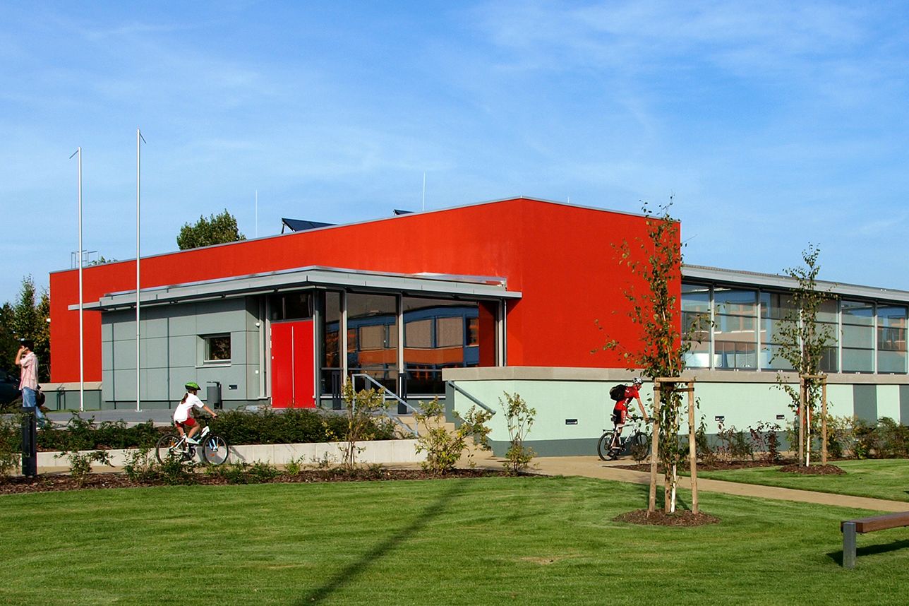 Mehrzweckhalle in den Farben rot und graugrün, davor eine grüne Wiese 