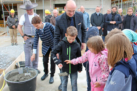 Grundsteinlegung für neue Kita Kinder und der Finanzbürgermeister verfüllen mit Zement das Loch, in dem die Grundsteinschatulle versenkt wurde. Im Hintergrund viele Menschen.