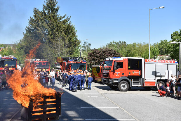 Tag der offenen Tür auf dem Gelände der Feuerwehr: im Vordergrund brennender Holzhaufen, im Hintergrund Feuerwehrfahrzeuge, davor Zuschauer und eine Gruppe der Jugendfeuerwehr bei der Übung das Feuer zu löschen