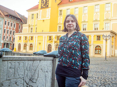Gleichstellungsbeauftragte Frau steht am Brunnen, im Hintergrund ein gelbes Haus