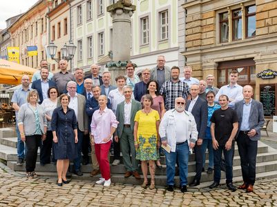 Gruppenfoto Stadträte Frauen und Männer stehen vor einem Brunnen, im Hintergrund farbige Häuserfassaden