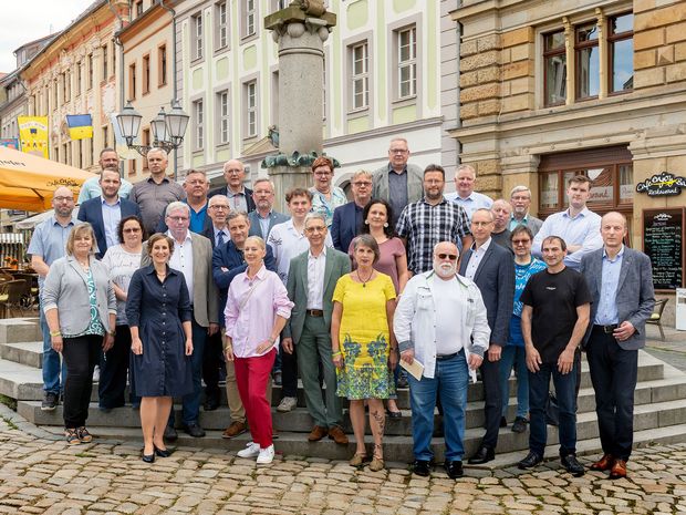 Frauen und Männer stehen vor einem Brunnen, im Hintergrund farbige Häuserfassaden