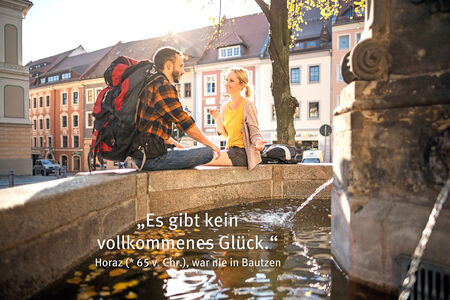 Vollkommenes Glück Junger Mann mit Rucksack und junge Frau sitzen auf Brunnenrand, im Hintergrund Häuserzeile