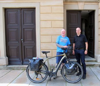 Oli radelt für einen guten Zweck Mann im blauen T-Shirt mit Fahrrad steht neben Mann in schwarzen T-Shirt und blauer Jeans vor einem hellgelben Gebäude mit brauner Tür