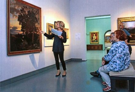 Frau im schwarzen Hosenanzug steht vor einem Ölgemälde und erläutert dies den Besuchern (zwei Männern und eine Frau).