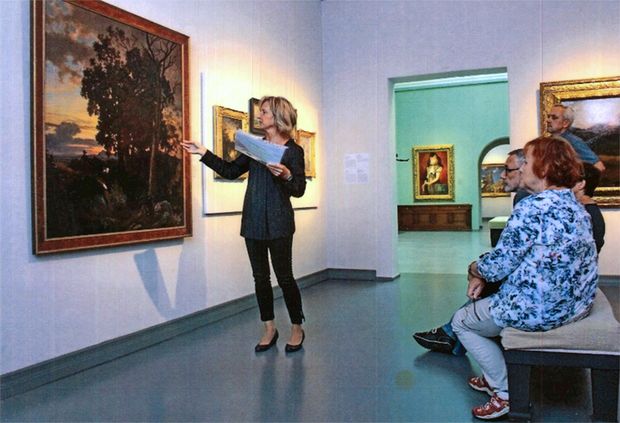 Frau im schwarzen Hosenanzug steht vor einem Ölgemälde und erläutert dies den Besuchern (zwei Männern und eine Frau).