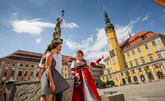Stadtführung mit Tuchhändlerin Teda Ein Paar – Mann und Frau stehen mit Tuchhändlerin Teda am Brunnen auf dem Hauptmarkt, im Hintergrund sieht man das Rathaus