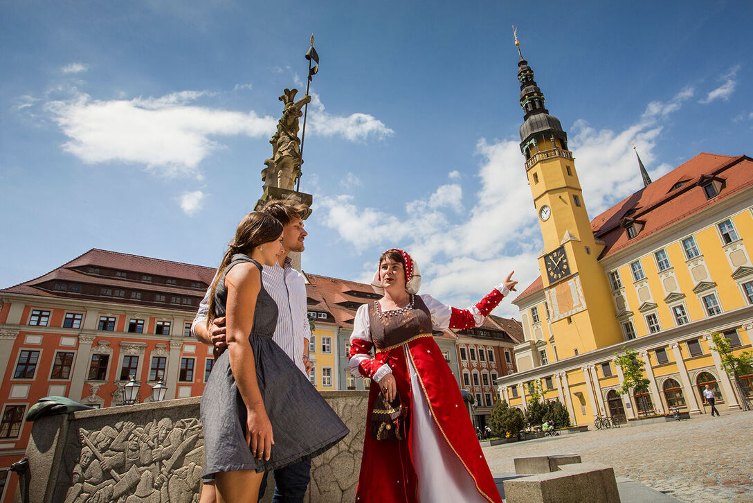 Stadtführung mit Tuchhändlerin Teda Ein Paar – Mann und Frau stehen mit Tuchhändlerin Teda am Brunnen auf dem Hauptmarkt, im Hintergrund sieht man das Rathaus