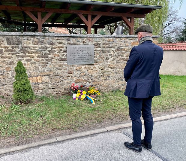 Mann in blauem Anzug steht vor Gedenktafel, darunter liegt eine Blumengebinde