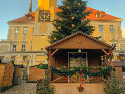 Elf Jarle auf der Bühne Bühne und Holzbuden mit Weihnachtsbaum, im Hintergrund das Rathaus, ein gelbes Gebäude mit Turm