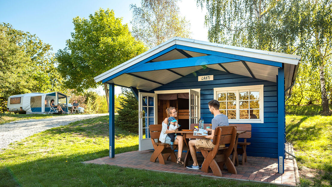 eine Familie sitzt unter dem Vordach einer Holzhütte an einem Tisch