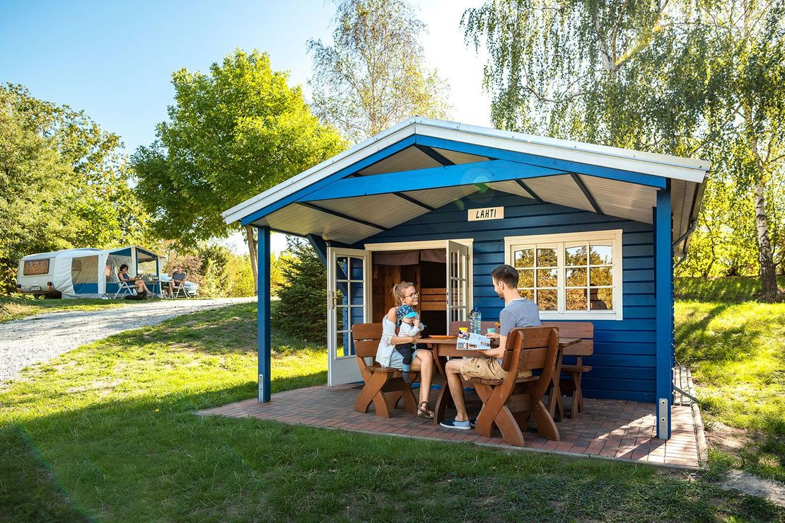 eine Familie sitzt unter dem Vordach einer Holzhütte an einem Tisch