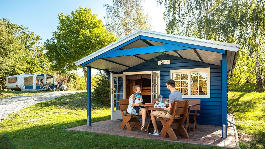 Stausee Campingplatz eine Familie sitzt unter dem Vordach einer Holzhütte an einem Tisch