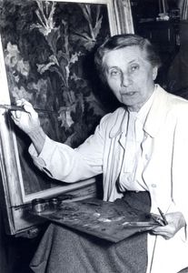 Marianne Britze schwarz-weiß Aufnahmen- eine Frau sitzt mit Farbpalette auf dem Schoß und Pinsel in der Hand vor ihrem Gemälde