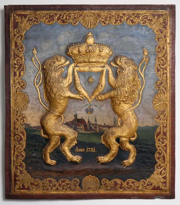 Ein rechteckiges Schild aus Metall. darauf wurde die Ansicht der Satdt Bautezn aus der ferne gemalt. Im Vordergrund stehen als Relief aufrecht zwei goldene Löwen, die in ihren Vorderpfoten eine goldene Krone über die Stadt halten. Es ist das Wappen der Tuchmacher aus 1731.