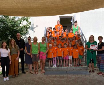 Auszeichnung Kita Kneipp Kinder in orangenen T-Shirts stehen auf einer Treppe links und rechts daneben stehen Erwachsene