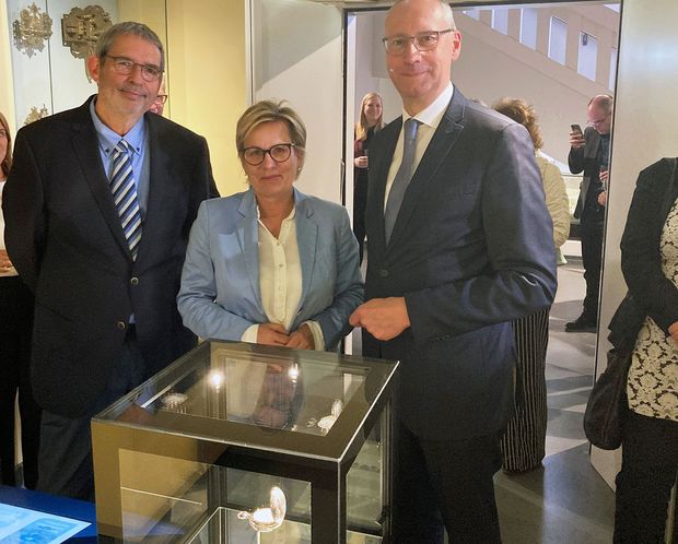 Tourbillon in Dauerausstellung Ein Mann, eine Frau und noch ein Mann stehen in dunkelblauen Anzügen gekleidet vor einer Glaskiste. Sie lächeln in die Kamera