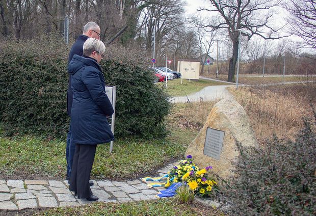 Frau in blauer Jacke und Mann in schwarzem Mantel stehen vor einen Gedenkstein. Davor liegen Blumengebinde.