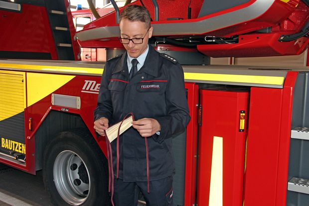Mann in Feuerwehrkleidung vor rotem Feuerwehrfahrzeug mit weißer Mundschutzmaske in der Hand