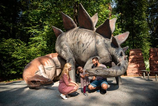 Saurierpark kleines Mädchen schaut mit Mutter und Vater ins Maul eines Stegosaurus. Daneben liegt ein Allosaurus.