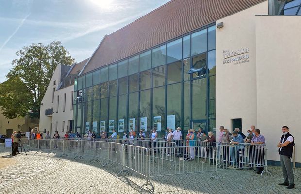 Eine Schlange Menschen steht hinter Zäunen vor einer riesigen Glaswand