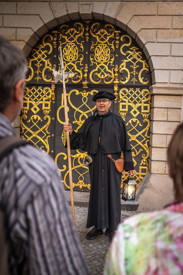 Stadtführung mit Nachtwächter Mann in historischem schwarzem Gewand steht vor einem goldverzierten Tor