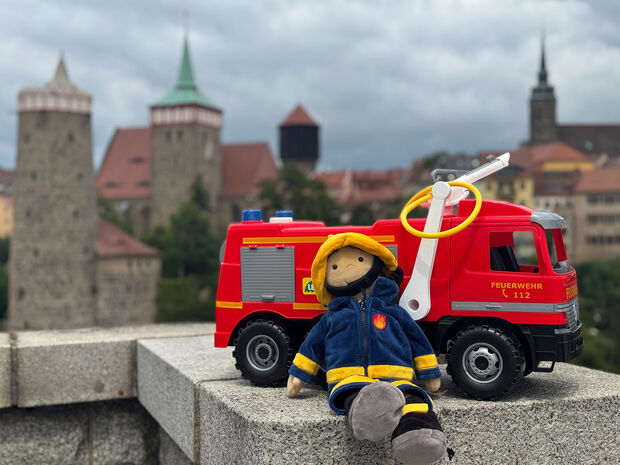 Maskottchen Oernie Feuerwehrmann als Plüschfigur (Kuscheltier) vor Spielzeug-Feuerwehrauto, im Hintergrund sieht man die Altstadt-Silhouette von Bautzen von der Friedensbrücke aus
