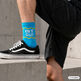 Mann mit kurzen schwarzen Hosen hat linken Fuß auf eine flache Mauer gestellt. Er trägt schwarze Sportschuhe mit weißer Sohle. Und türkisfarbene Socken mit weißer Schrift am Schaft Ist alles Wurst