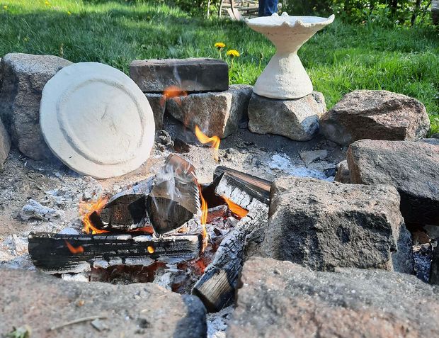 Foto einer offenen Feuerstelle in einer runden Steinsetzung und ein vergrößerter Nachbau eines eisenzeitlichen Ofenmodells aus Niederkaina aus Ton vor dem Brand.